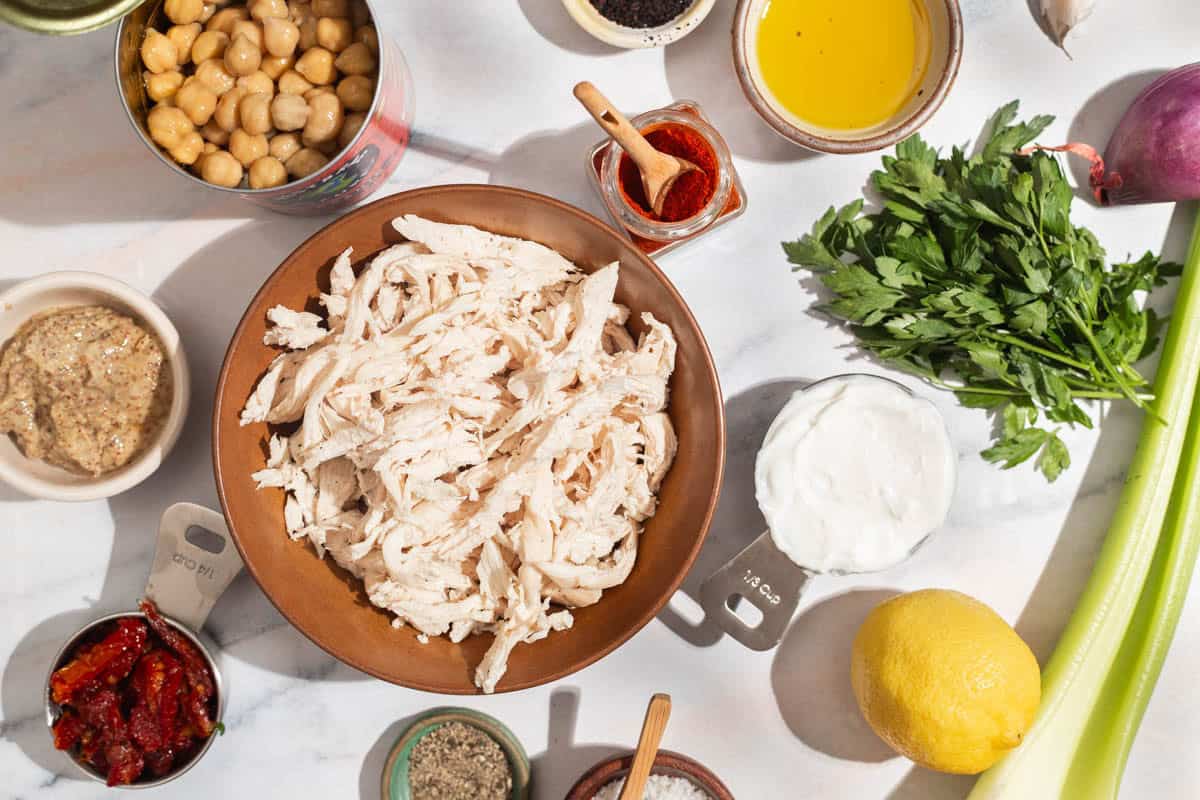 Ingredients for chickpea chicken salad including shredded chicken breast, sun dried tomatoes, chickpeas, greek yogurt, dijon mustard, lemon, garlic, olive oil, paprika, urfa biber, salt, black pepper, celery, and shallots.