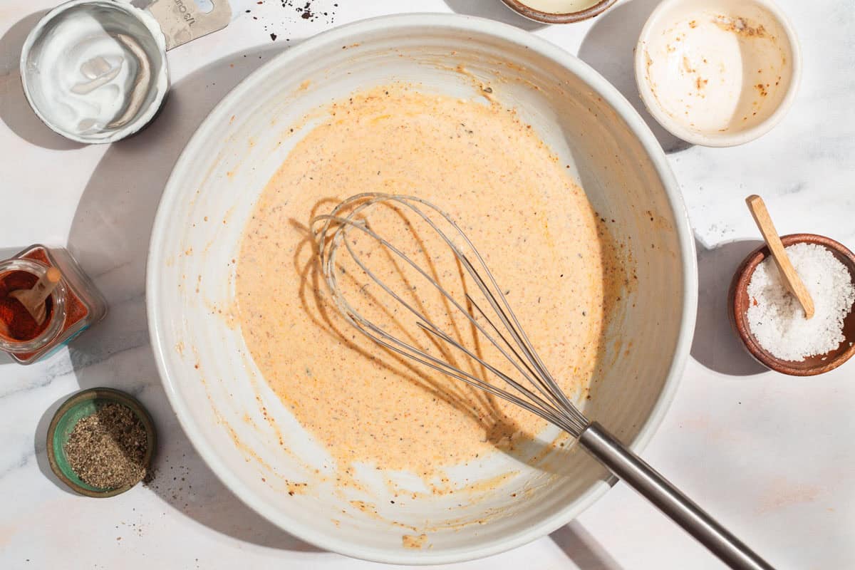 Yogurt, mustard, lemon juice, olive oil and spices mixed together in a bowl with a whisk. Surrounding this an empty bowl, an empty measuring cup, bowls of salt and pepper, and a jar of paprika.