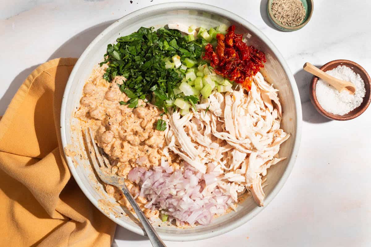 An overhead photo of the ingredients for the chickpea chicken salad in a bowl with a fork, just before being mixed together. Next to this is a cloth napkin and small bowls of salt and pepper.