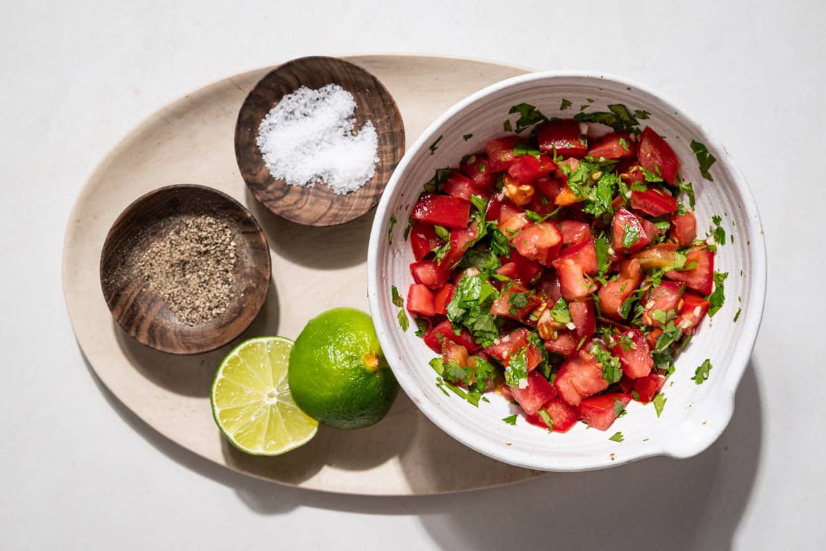 An overhead photo of the bruschetta topping a bowl on a platter with 2 lime halves and small bowls of salt and pepper.