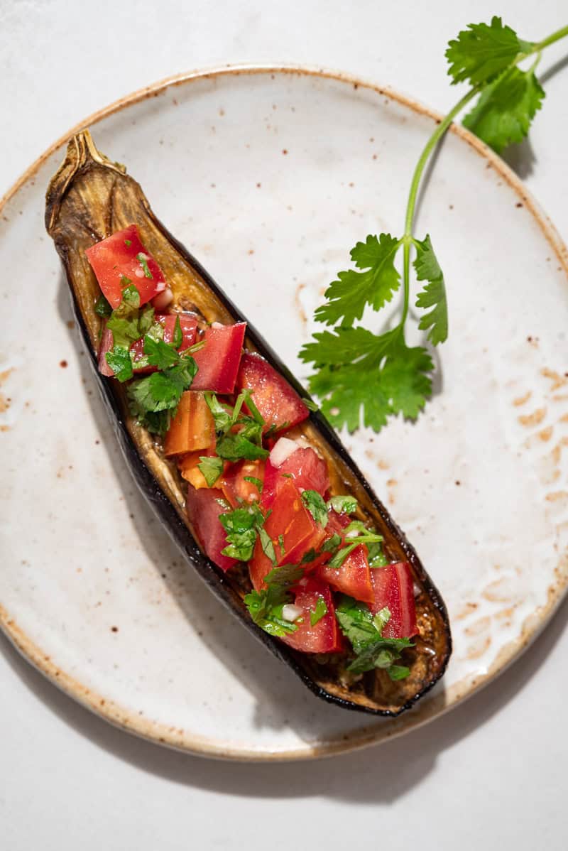 An overhead photo of 1 piece of eggplant bruschetta on a plate next to a sprig of cilantro.