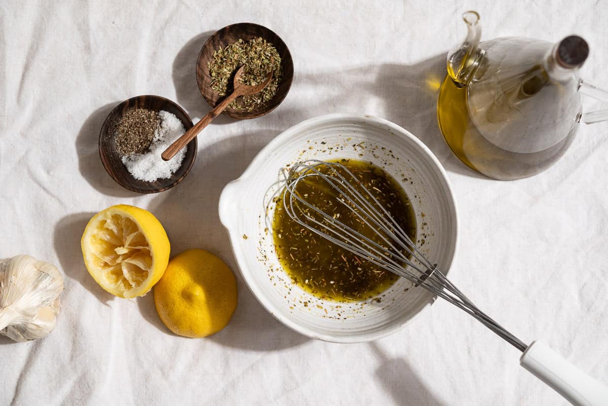 An overhead photo of the dressing for the eggplant salad in a bowl with a whisk. Next to this is a head of garlic, 2 juiced lemon halves, a bottle of olive oil, and bowl of salt and pepper and a bowl of za'atar.