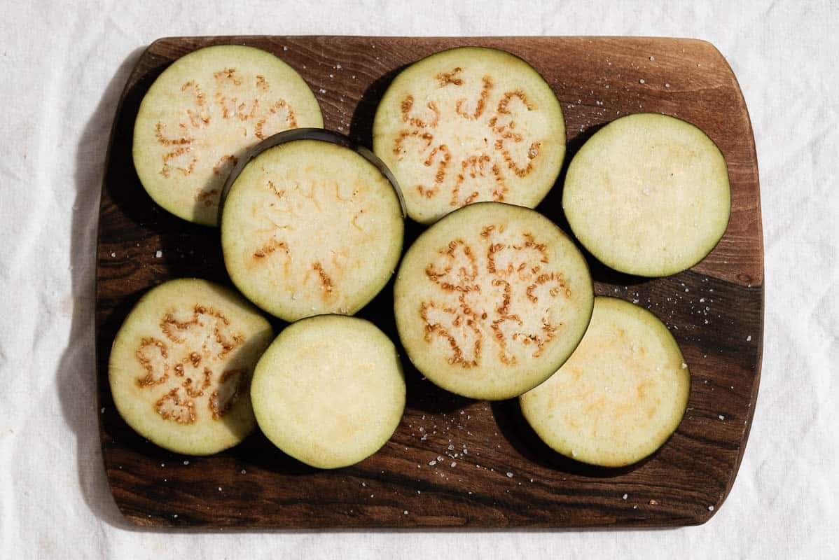 An overhead photo of sliced and salted eggplant on a wooden cutting board.