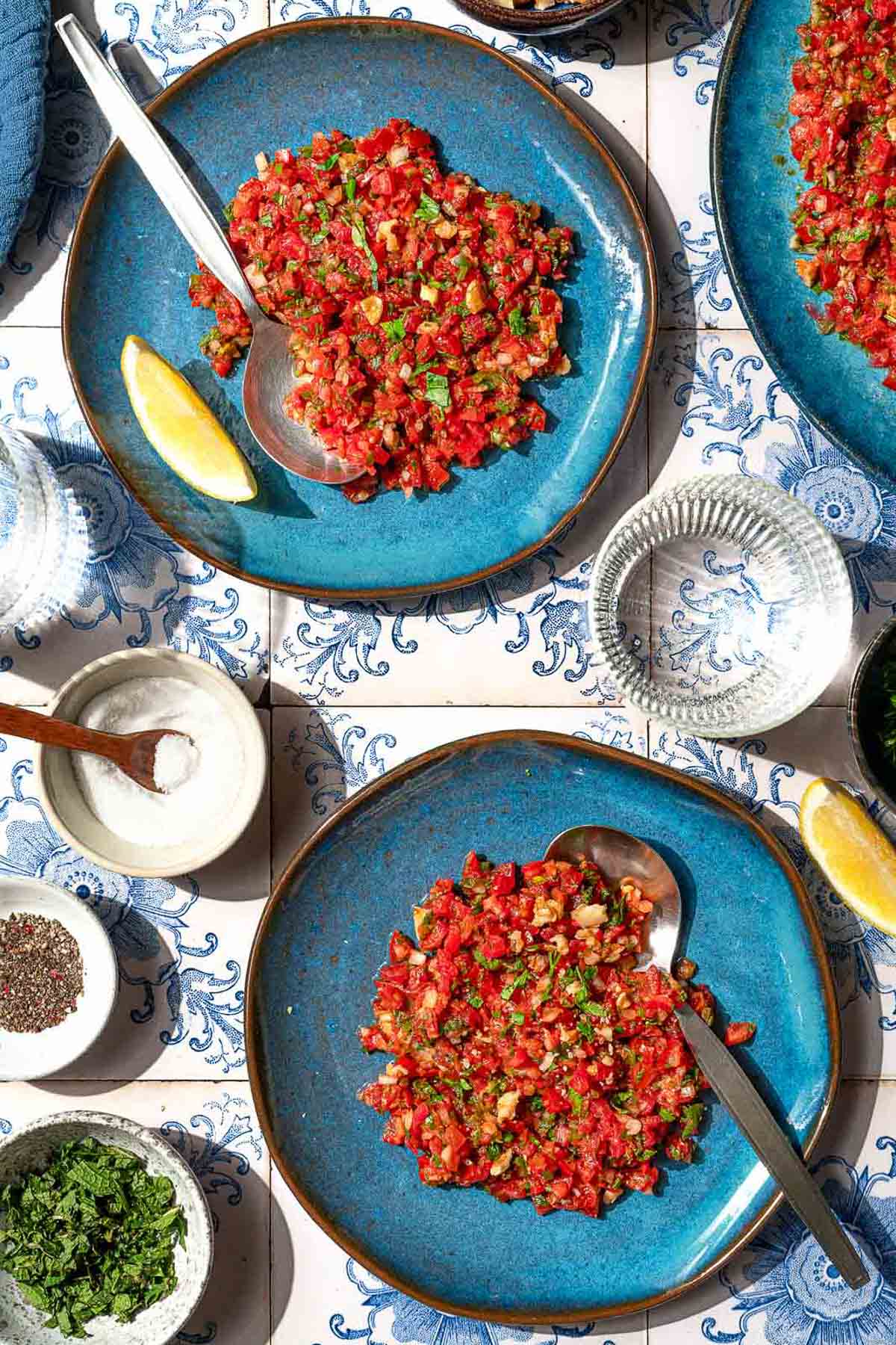 An overhead photo of 2 servings of ezme turkish salsa on plates with spoons. Next to these are bowls of salt, spices, and mint, lemon wedges, a glass of water, and a platter with the rest of the ezme.