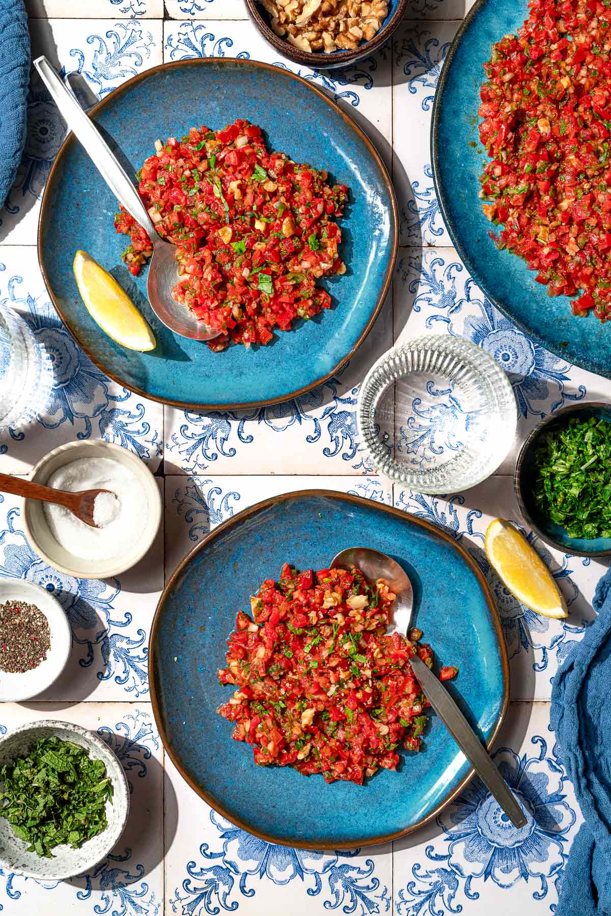 An overhead photo of 2 servings of ezme turkish salsa on plates with spoons. Next to these are bowls of salt, spices, mint and parsley, lemon wedges, a glass of water, and a platter with the rest of the ezme.