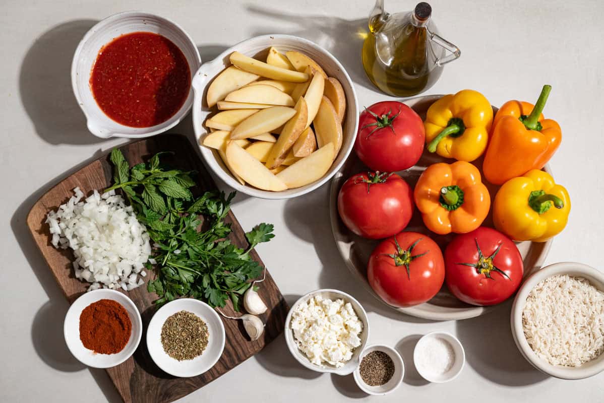 Ingredients for gemista greek stuffed vegetables including bell peppers, tomatoes, potatoes, rice, yellow onion, garlic cloves, parsley, mint, oregano, paprika, feta, olive oil, crushed tomatoes, salt and ground pepper.