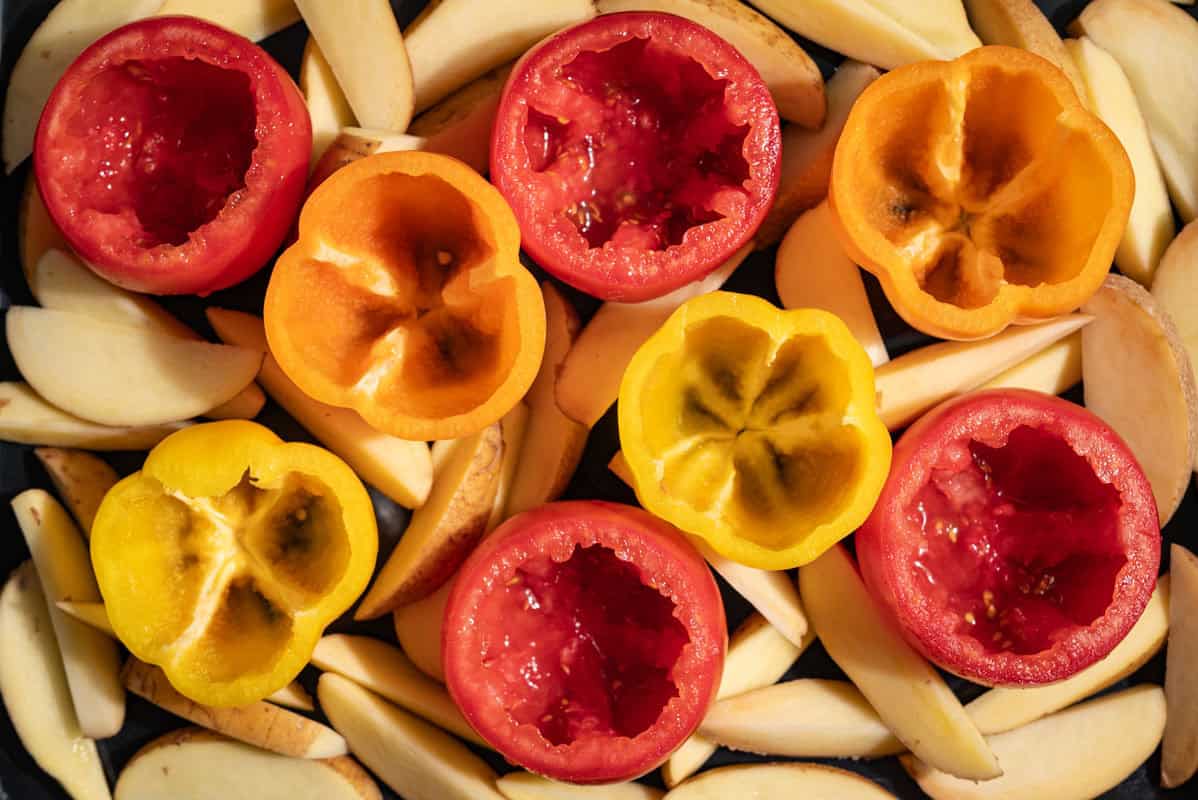 An overhead close up photo of bell peppers and tomato halve hollowed out in a backing dish surrounded by unbaked potato wedges.