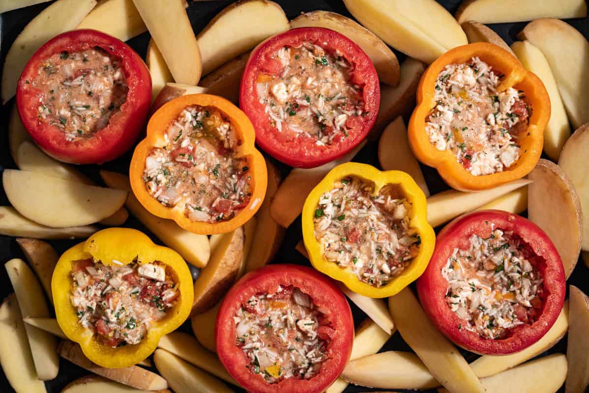 An overhead close up photo of bell peppers and tomato halves stuffed with the rice filling in a backing dish surrounded by unbaked potato wedges.