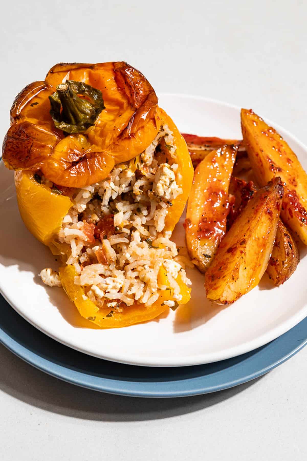 A close up of a stuffed yellow pepper with a serving of potato wedges on a stack of 2 plates.