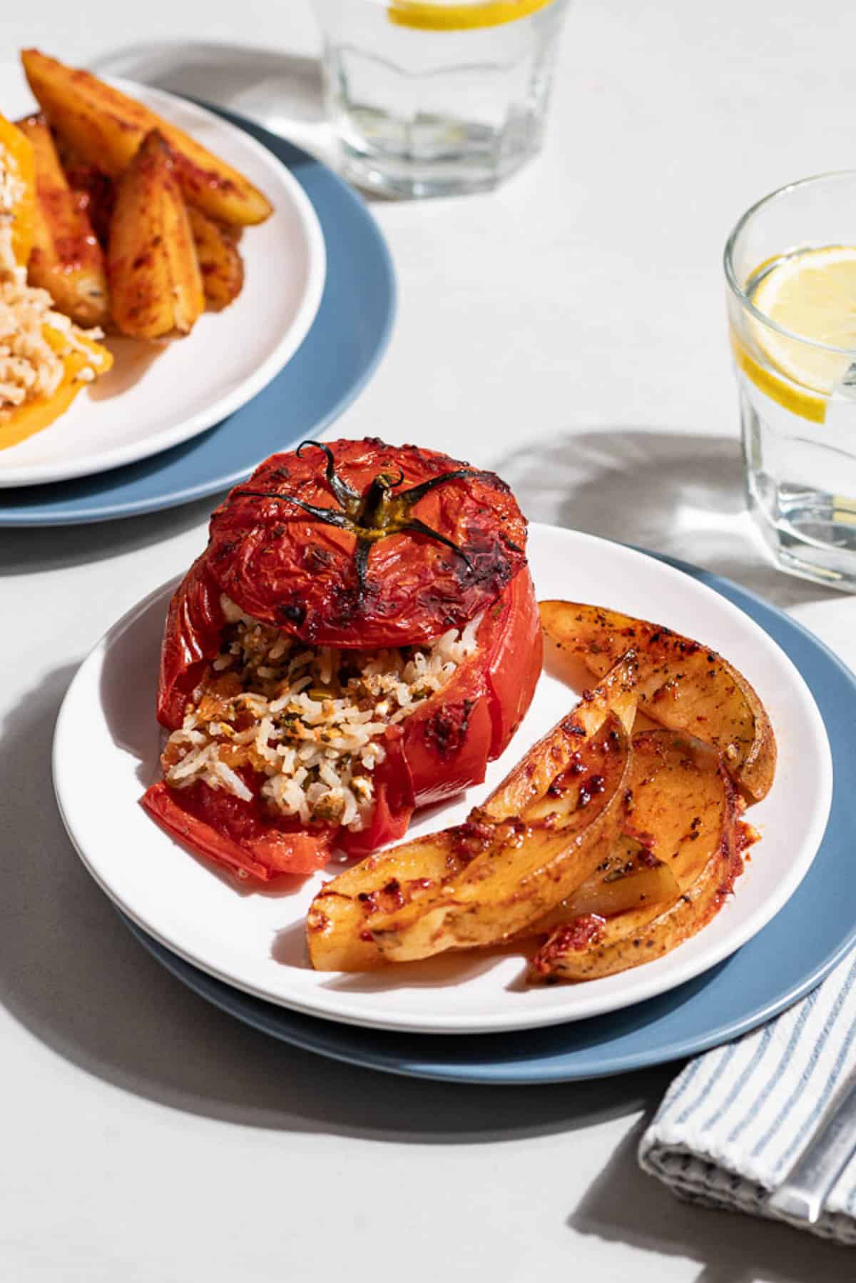 A close up of a stuffed tomato with a serving of potato wedges on a stack of 2 plates. Next to this is another serving of the gemista and potatoes on a stack of 2 plates, two glasses of water with lemon wedges and a cloth napkin.