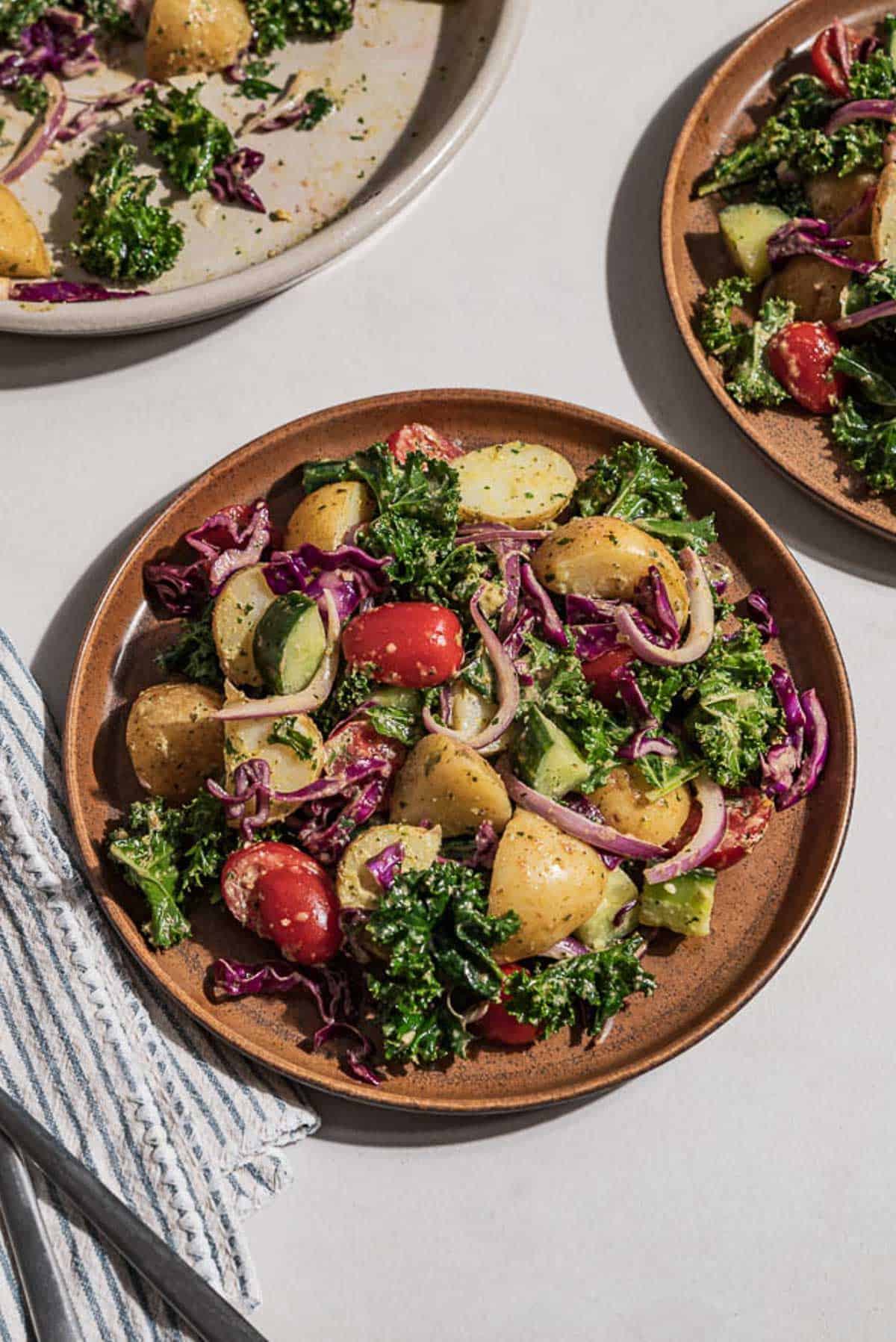 An overhead photo of vegetable laden potato salad on a plate next to a cloth napkin with a fork, another plate of the salad, and a serving bowl with the salad.