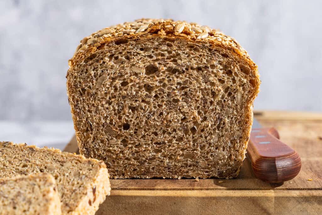 A close up photo of a partially sliced whole wheat bread loaf on a cutting board with a knife.