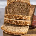 A close up photo of a partially sliced whole wheat bread loaf behind 3 slices of the bread on a cutting board with a knife.