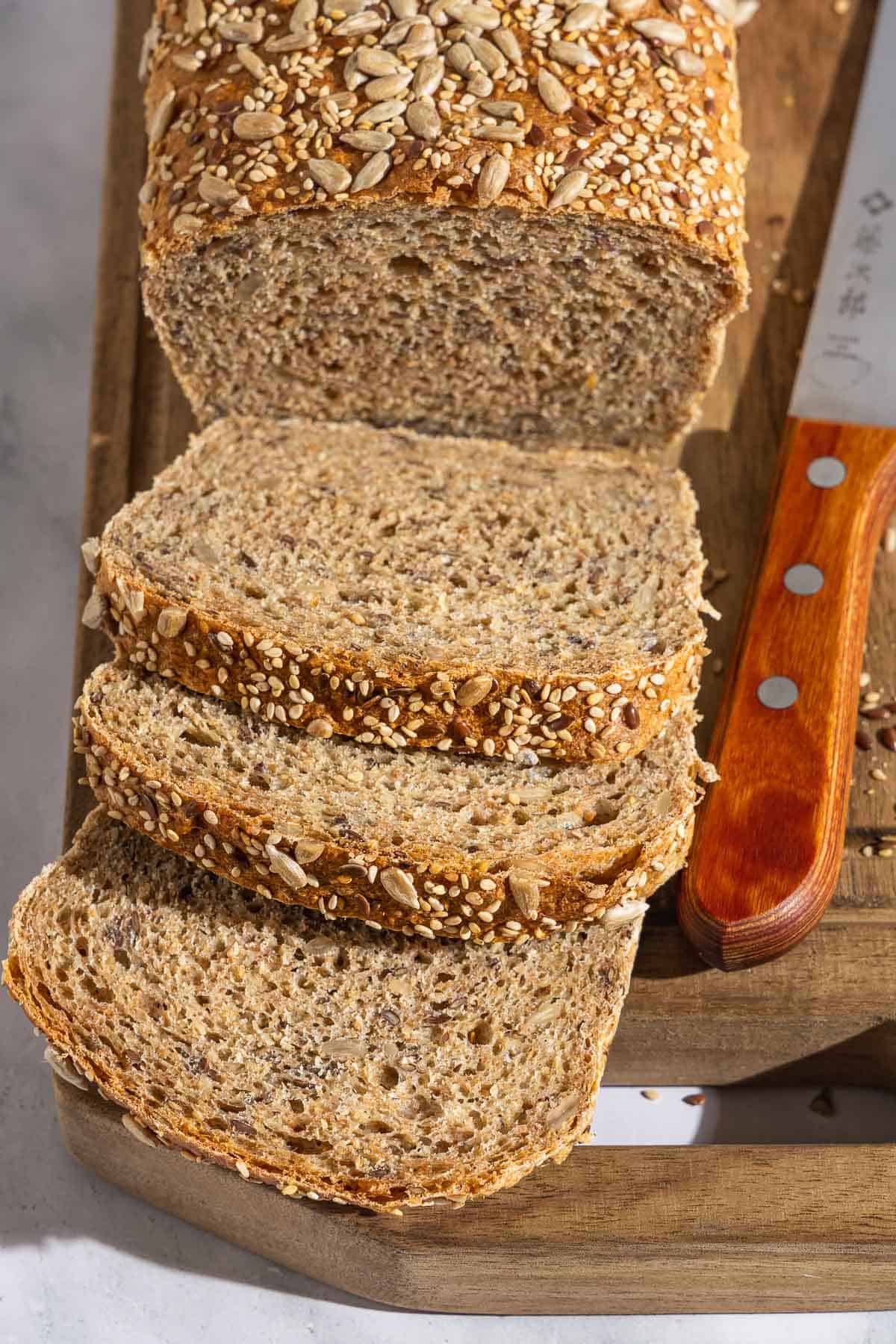 An overhead photo of a partially sliced whole wheat bread loaf behind 3 slices of the bread on a cutting board with a knife.