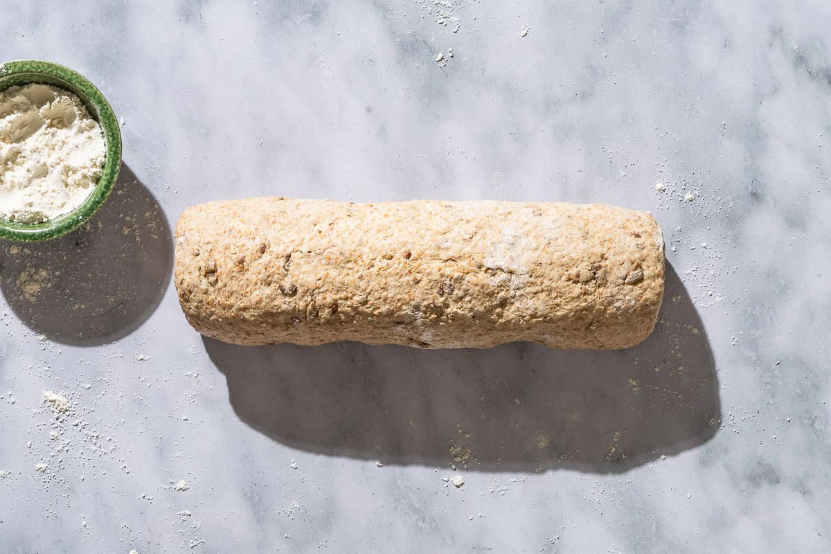 An overhead photo of whole wheat bread dough rolled into a cylinder shape next to a small bowl of flour.