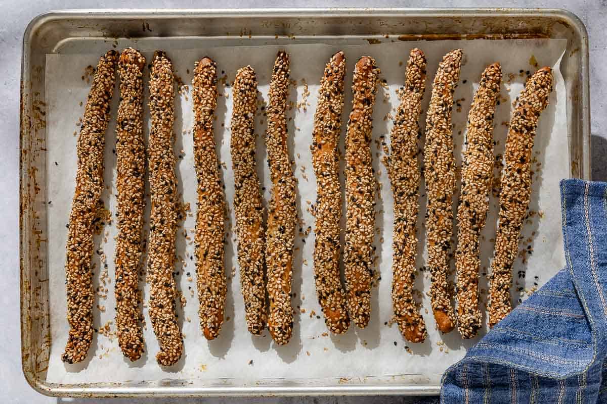 An overhead photo of 12 baked crispy sesame breadsticks on a parchment lined sheet pan next to a kitchen towel.