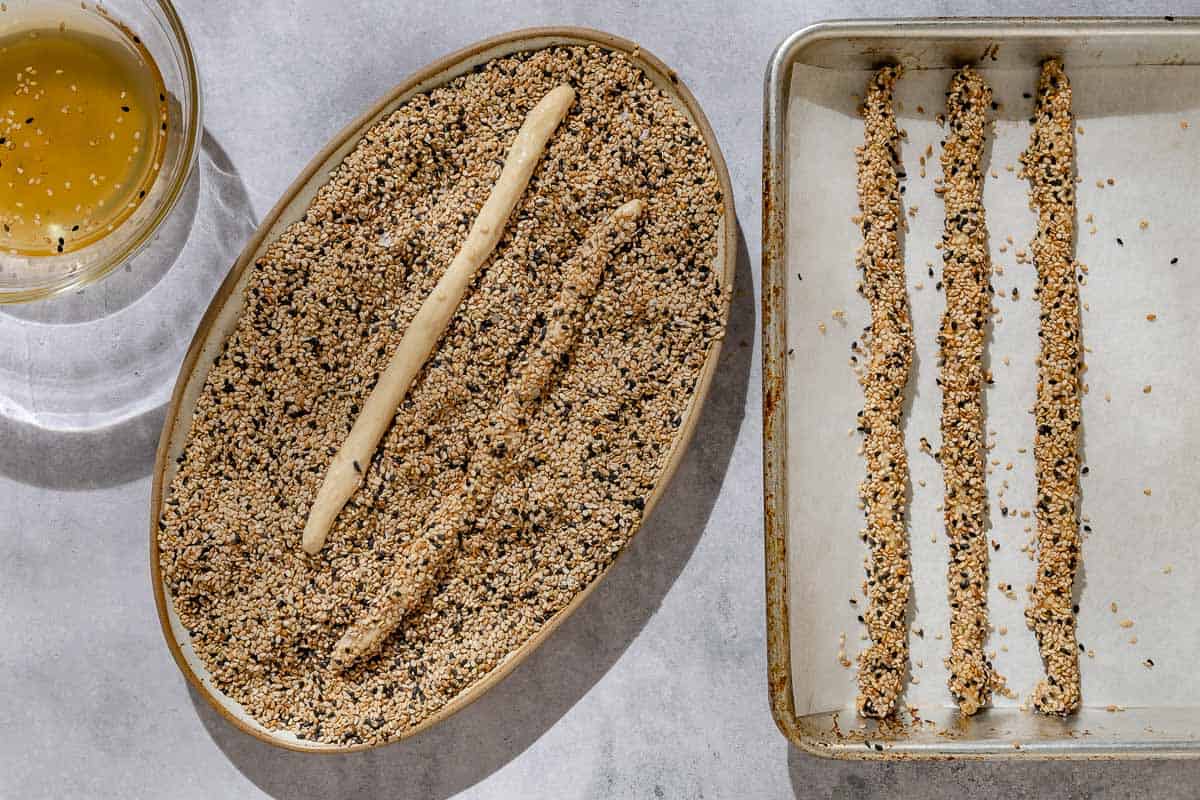 An overhead photo of 2 ropes of dough being rolled in sesame seeds in a large bowl, next to a bowl of honey water, and 3 unbaked sesame breadsticks on a parchment lined sheet pan.