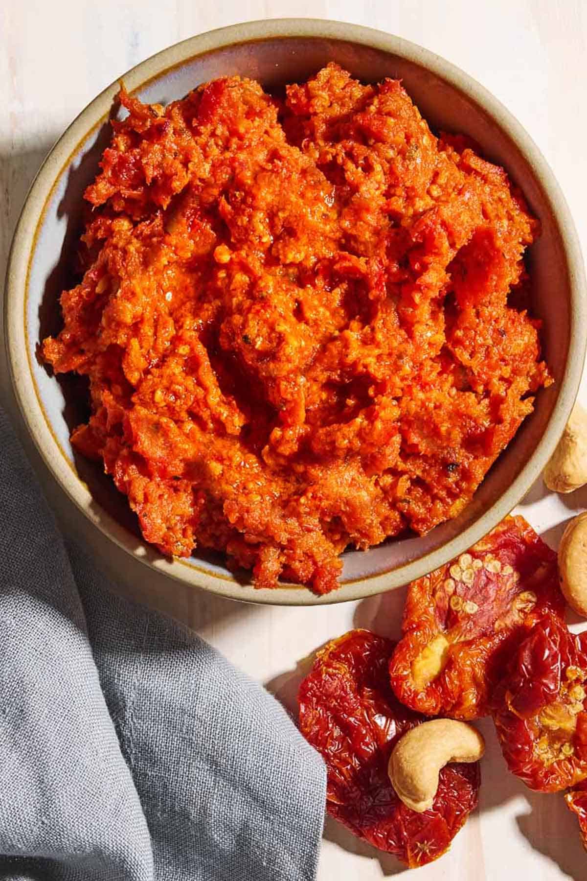 An overhead photo of sun dried tomato pesto in a bowl. Next to this is a cloth napkin, and some cashews and sun dried tomatoes.