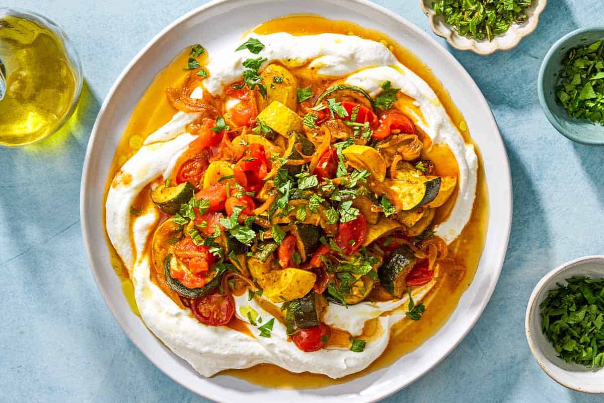 An overhead photo of borani kadoo on a serving plate. Next to this is a bottle of olive oil and 3 small bowls of chopped herbs.