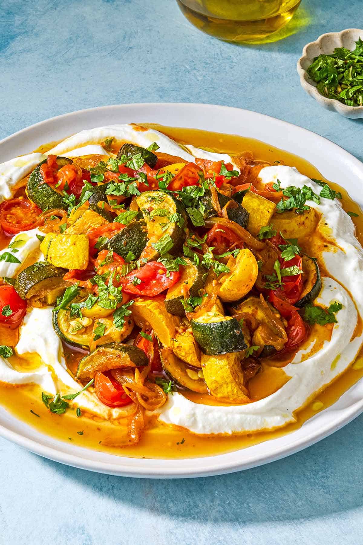 Borani kadoo on a serving plate. Next to this is a small bowl of chopped herbs and a bottle of olive oil.