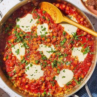 An overhead photo of chickpeas in tomato sauce with a wooden spoon in a skillet. Next this is a plate with crusty bread, a kitchen towel and a small bowl of pecorino romano cheese.