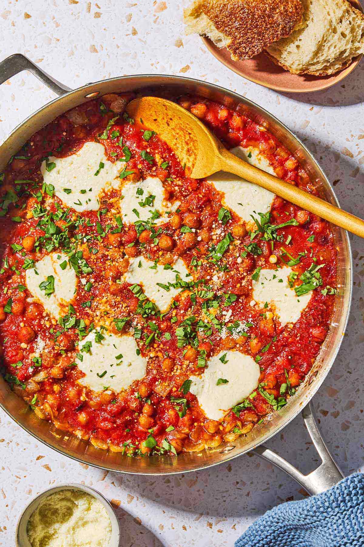 An overhead photo of chickpeas in tomato sauce with a wooden spoon in a skillet. Next this is a plate with crusty bread, a kitchen towel and a small bowl of pecorino romano cheese.