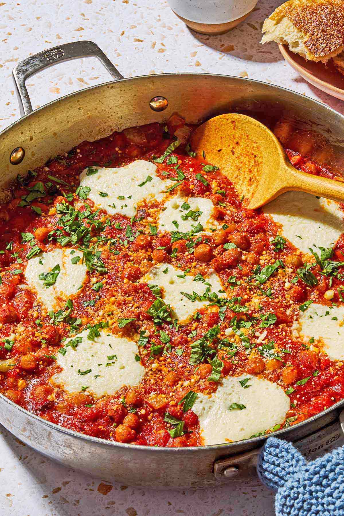 A close up photo of chickpeas in tomato sauce with a wooden spoon in a skillet. Next this is a plate with crusty bread.