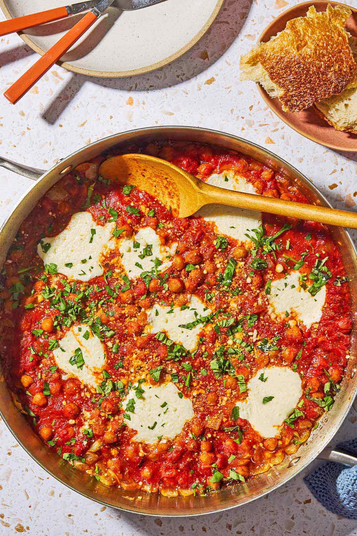 An overhead photo of chickpeas in tomato sauce with a wooden spoon in a skillet. Next this is a plate with utensils, and a plate with some crusty bread.