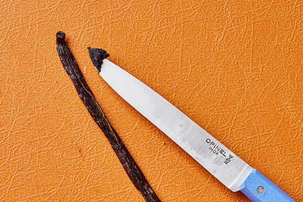 A close up of a vanilla bean and a knife with a bit of the vanilla bean seeds on it.