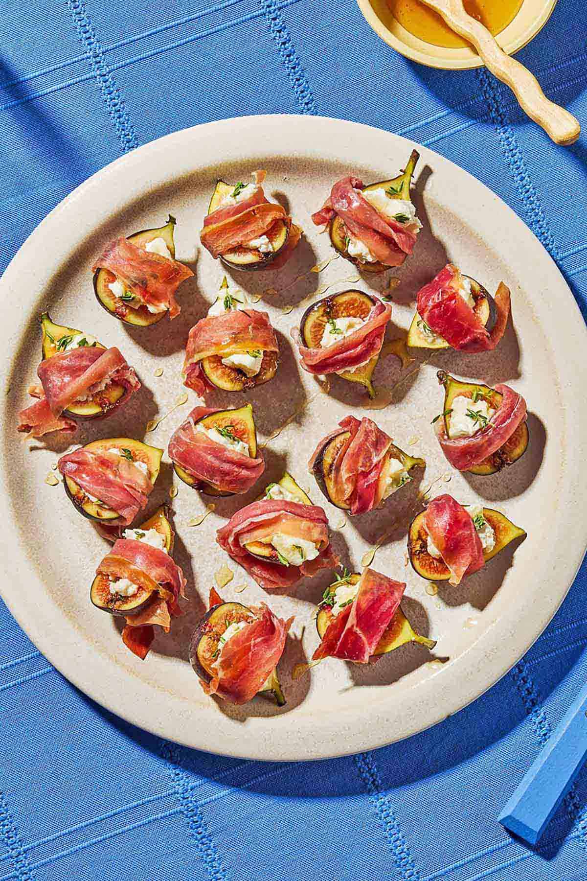 An overhead photo of several prosciutto wrapped figs drizzled with honey on a plate next to a bowl of honey.