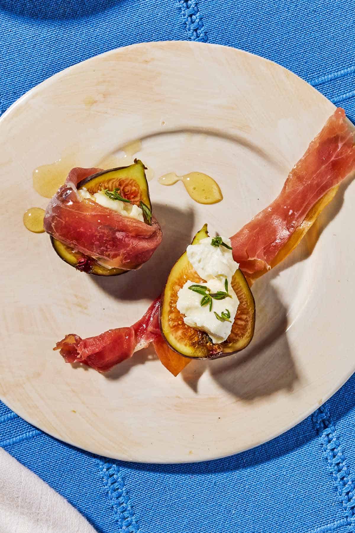 An overhead photo 2 fig halves topped with goat cheese and thyme on a plate. One is wrapped in prosciutto and one is sitting on a slice of prosciutto.