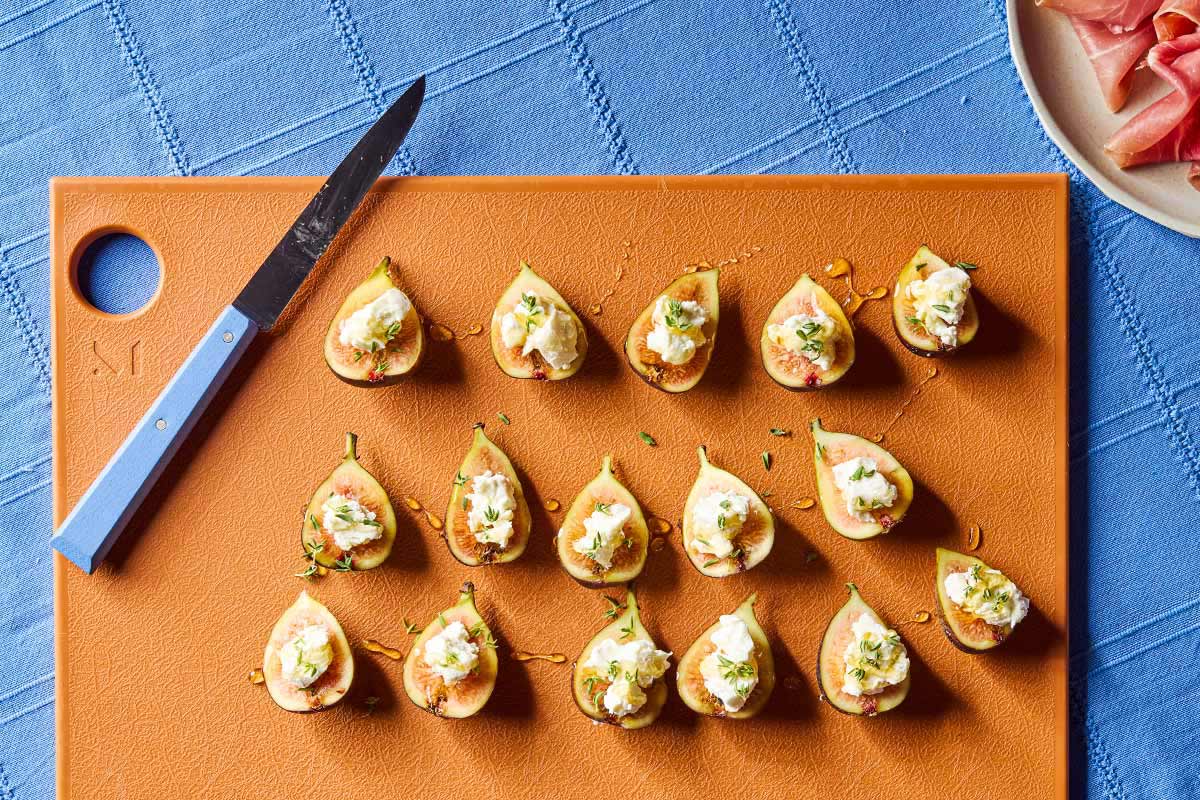 An overhead photo of several fig halves topped with goat cheese and thyme on a cutting board with a knife. Next to this is a plate of prosciutto.