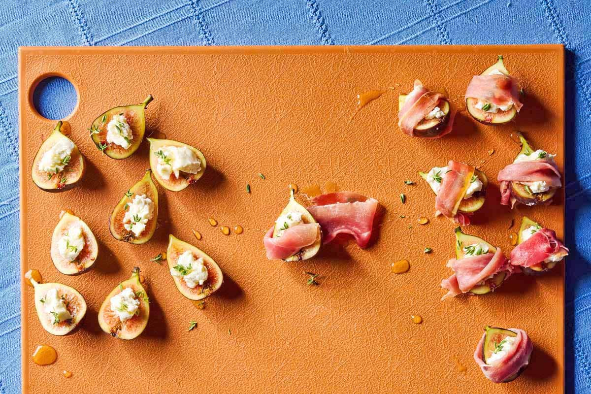 An overhead photo of several fig halves topped with goat cheese and thyme on a cutting board. Half of them are wrapped in prosciutto.