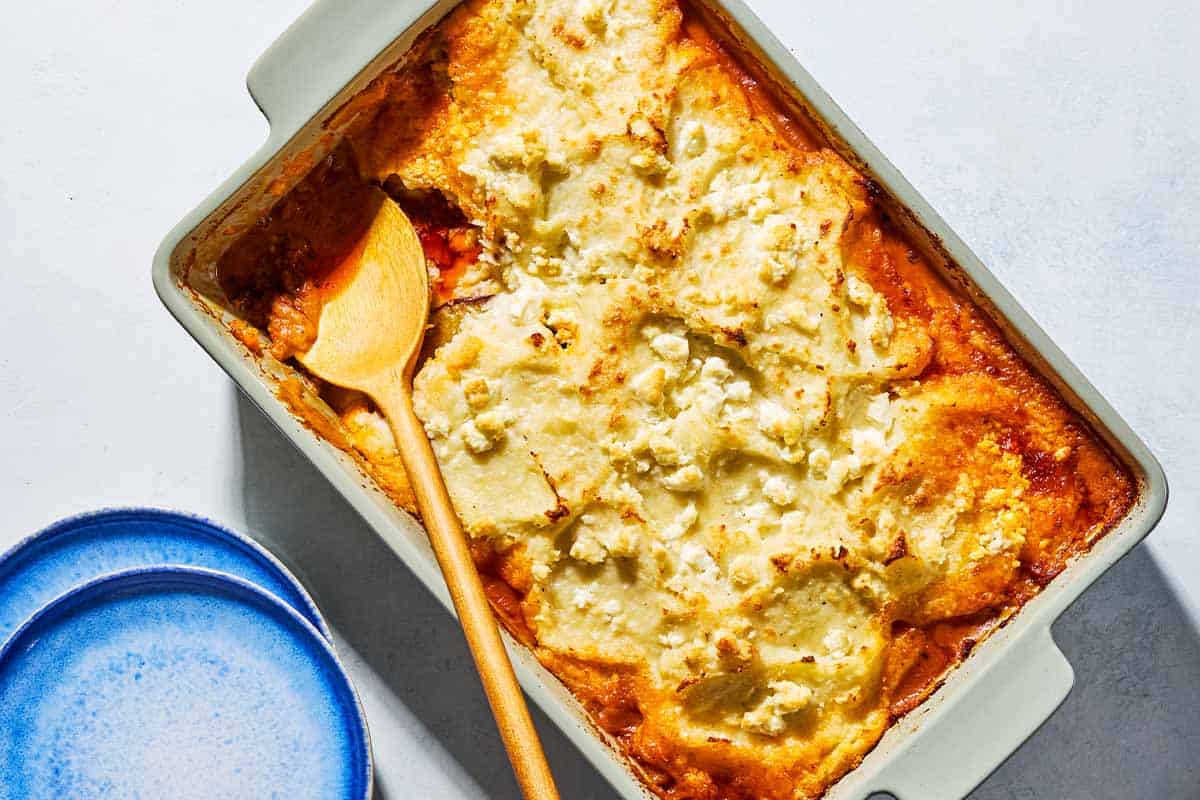 An overhead photo of Greek Shepherd's Pie in a baking dish with a wooden spoon. Next to this is a stack of 2 plates.