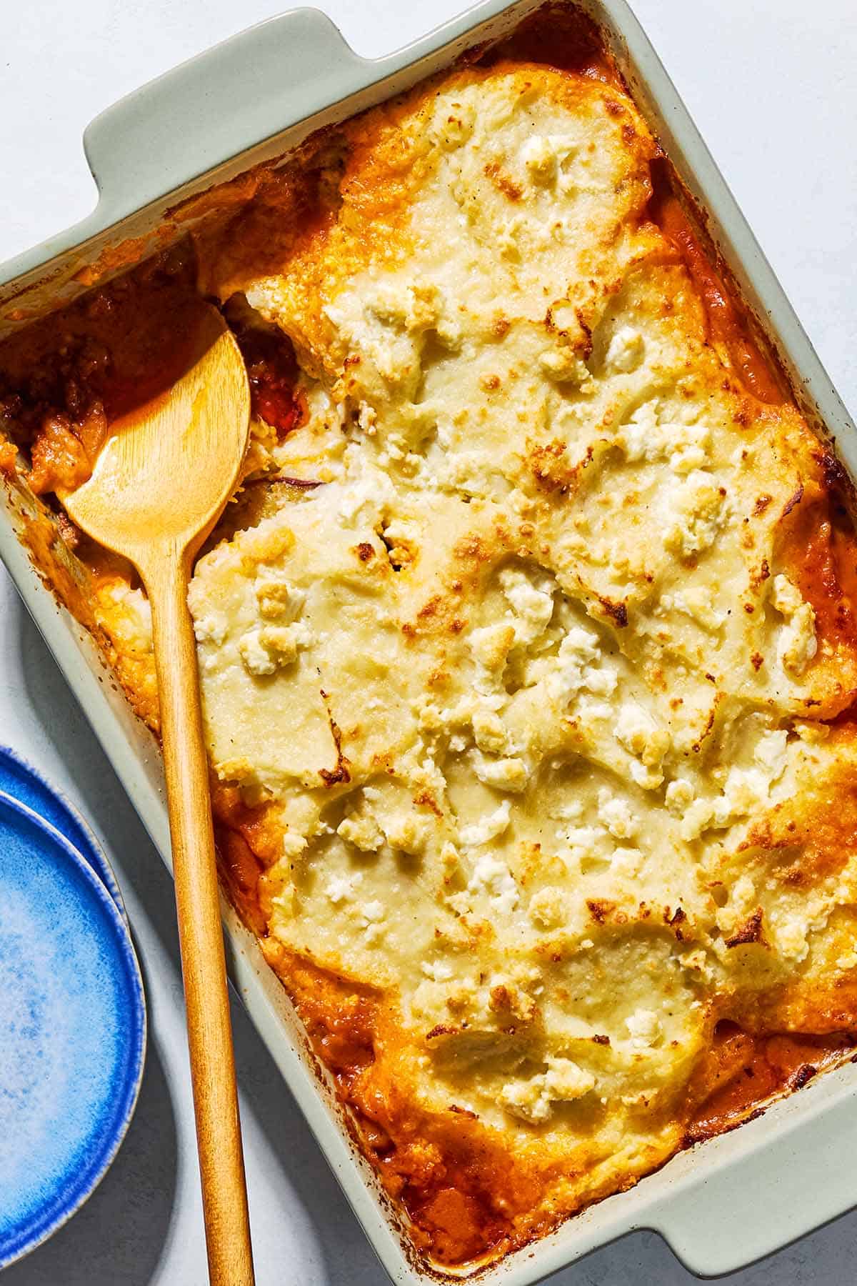 An overhead photo of Greek Shepherd's Pie in a baking dish with a wooden spoon. Next to this is a stack of 2 plates.