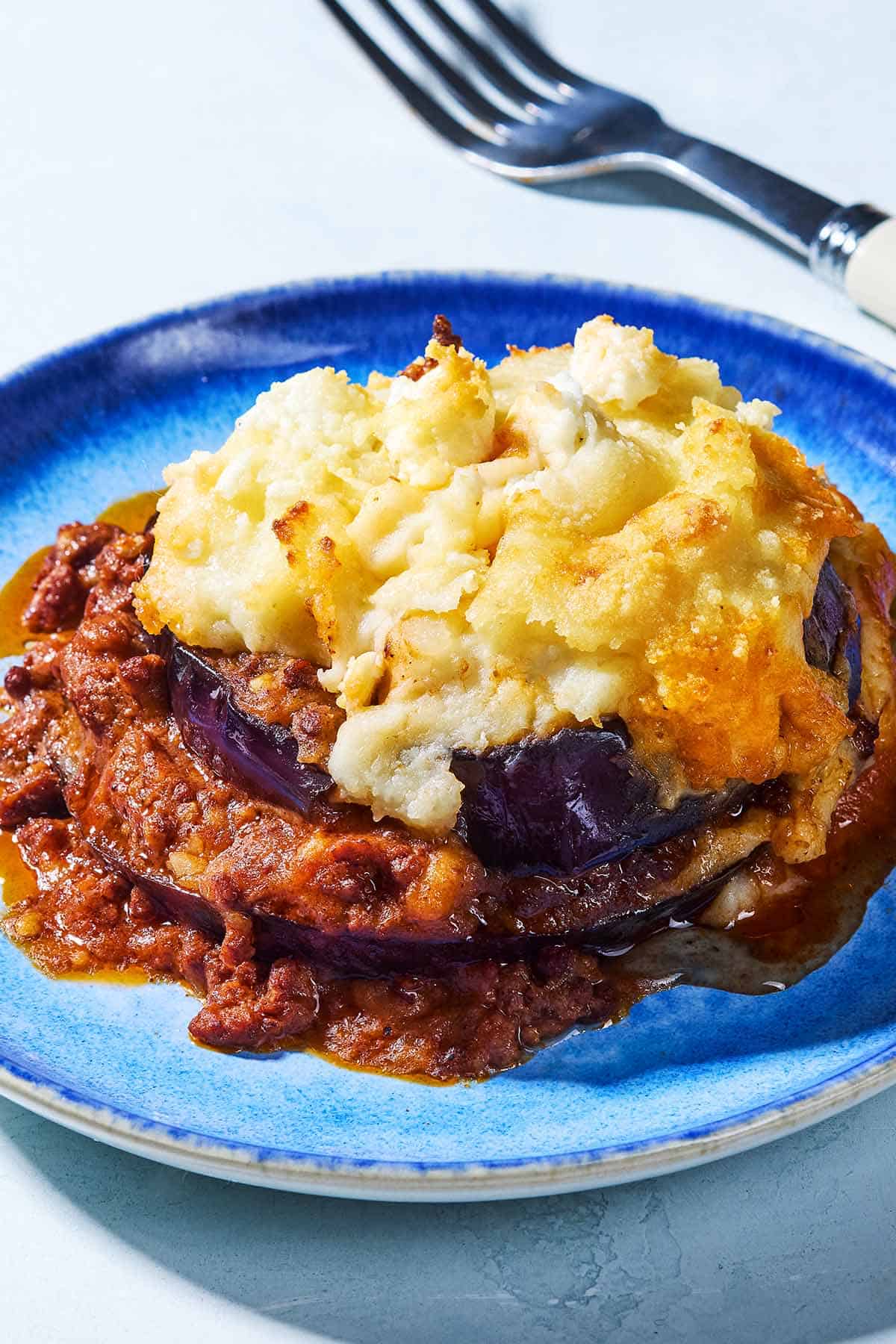 A close up of a serving of Greek Shepherd's Pie on a plate next to a fork.