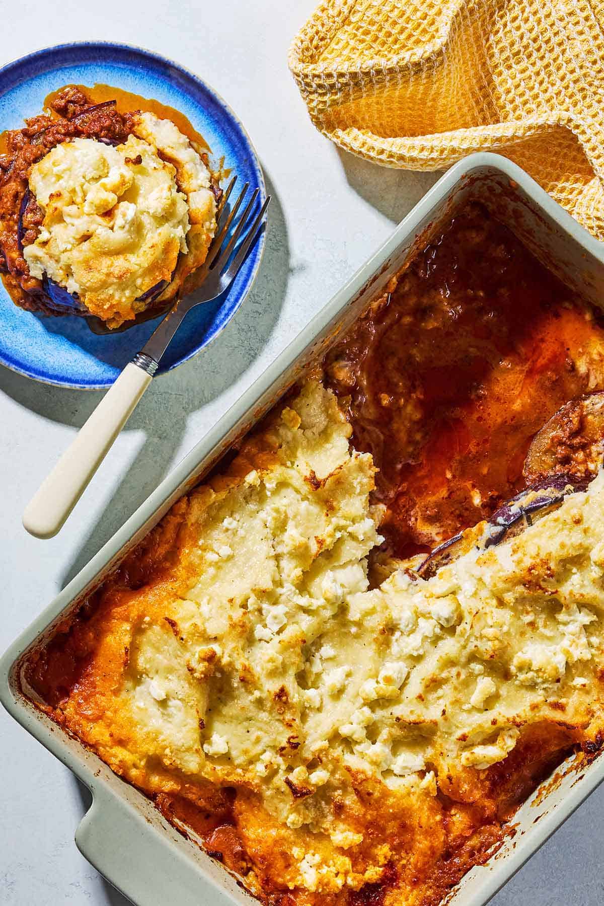 An overhead photo of Greek Shepherd's Pie in a baking dish with a few servings removed. Next to this is a kitchen towel and a serving of the pie on a plate with a fork.