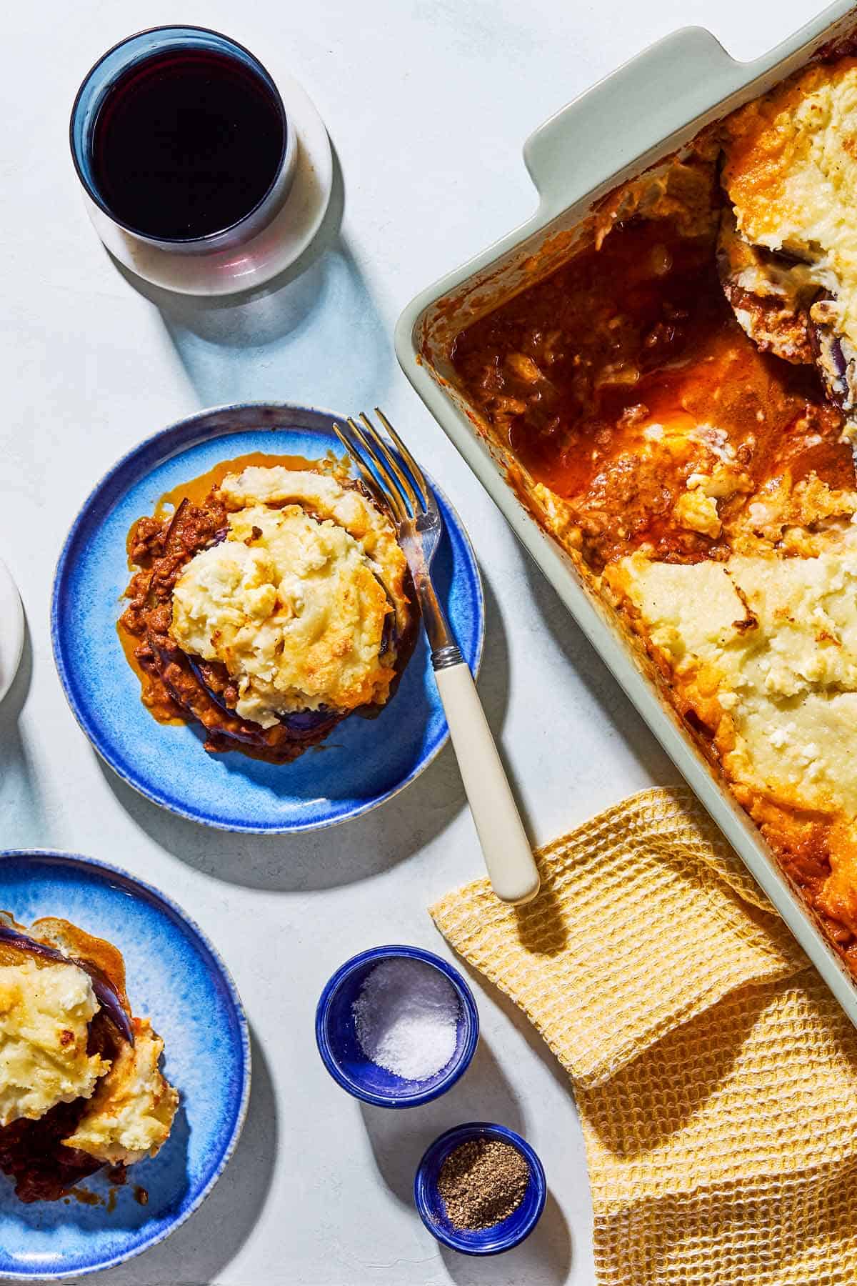 A closeup, overhead photo of Greek Shepherd's Pie in a baking dish with a few servings removed. Next to this is a kitchen towel, two servings of the pie on plates with forks, small bowels of salt and pepper, and a glass of red wine.