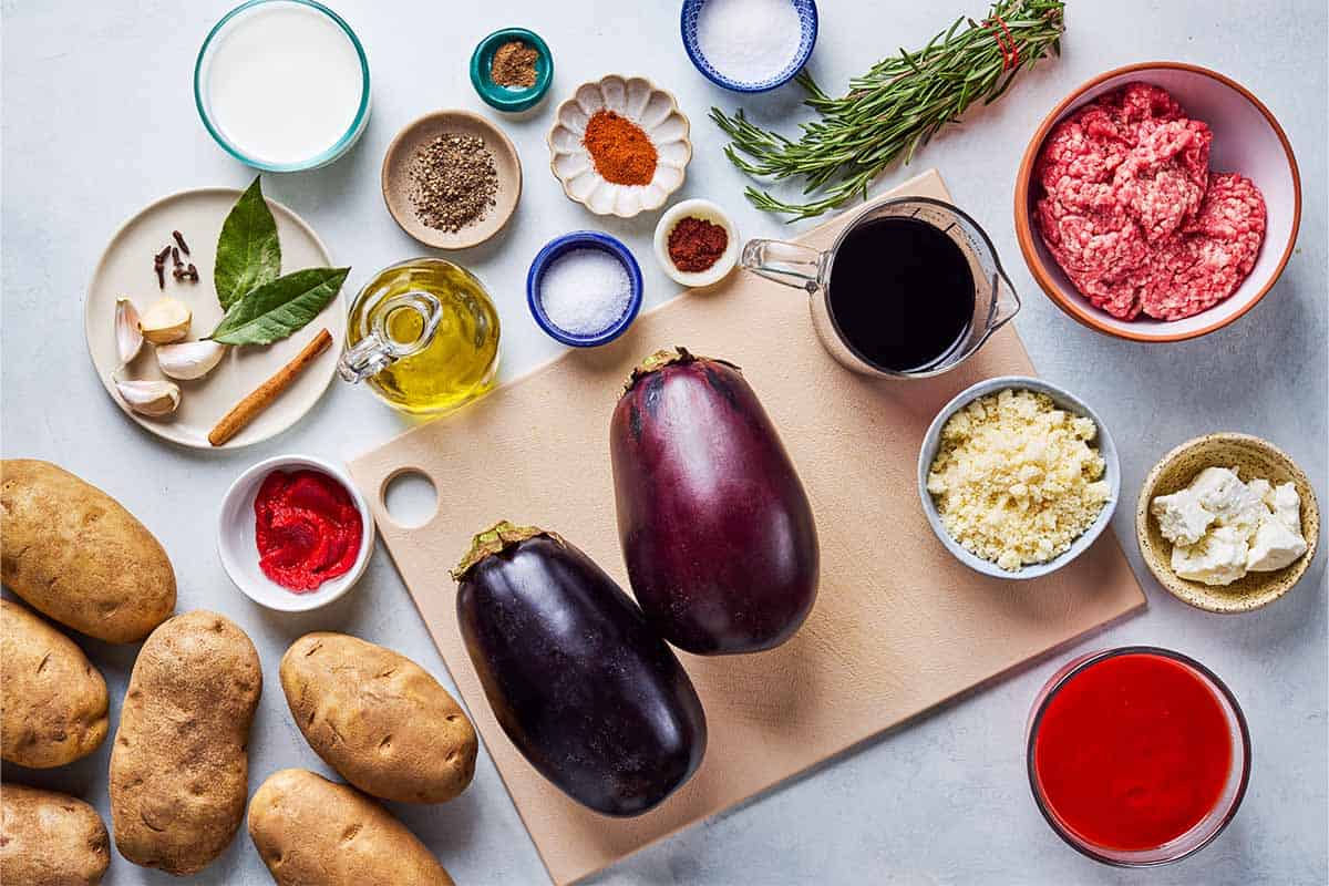 Ingredients for Greek Shepherd's Pie including olive oil, garlic, ground beef, salt, black pepper, sweet paprika, cinnamon, ground clove, tomato paste, tomato puree, red wine, water, cinnamon stick, whole cloves, sugar, globe eggplants, chopped fresh rosemary, russet potatoes, milk, grate parmesan, and crumbled feta.