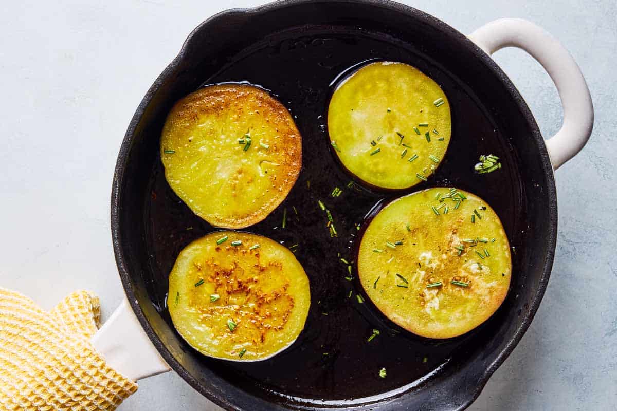 An overhead photo of 4 slices of eggplant being fried in a skillet. A kitchen towel is wrapped around the handle.