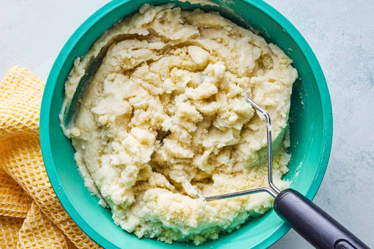 An overhead photo of the potato mash in a mixing bowl with a potato masher. Next to this is a kitchen towel.