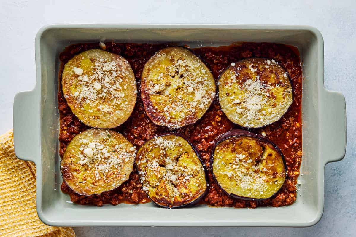 An overhead photo of the meat and eggplant layers of the Greek shepherd pi in a baking dish next to a kitchen towel.