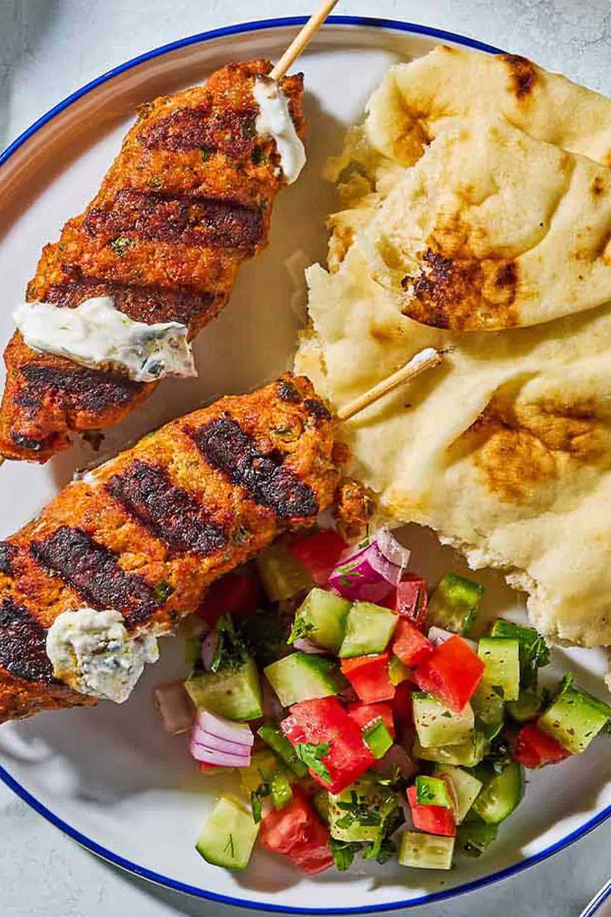 A close up of 2 grilled chicken kofta kebabs topped with mint yogurt sauce on a plate with pieces of pita and a tomato, cucumber salad.