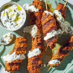 Six grilled chicken kofta kebabs topped with mint yogurt sauce on a plate with the rest of the sauce in a bowl, and spoon.