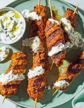 An overhead photo of six grilled chicken kofta kebabs topped with mint yogurt sauce on a plate with the rest of the sauce in a bowl, and spoon. Next to this is a cloth napkin.