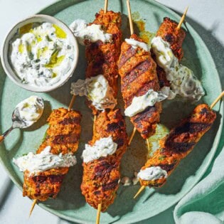 An overhead photo of six grilled chicken kofta kebabs topped with mint yogurt sauce on a plate with the rest of the sauce in a bowl, and spoon. Next to this is a cloth napkin.