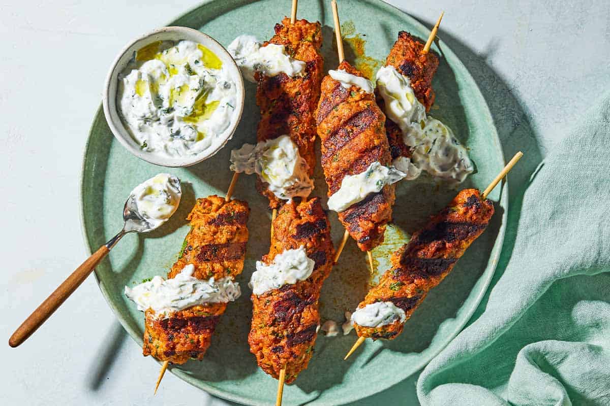 An overhead photo of six grilled chicken kofta kebabs topped with mint yogurt sauce on a plate with the rest of the sauce in a bowl, and spoon. Next to this is a cloth napkin.