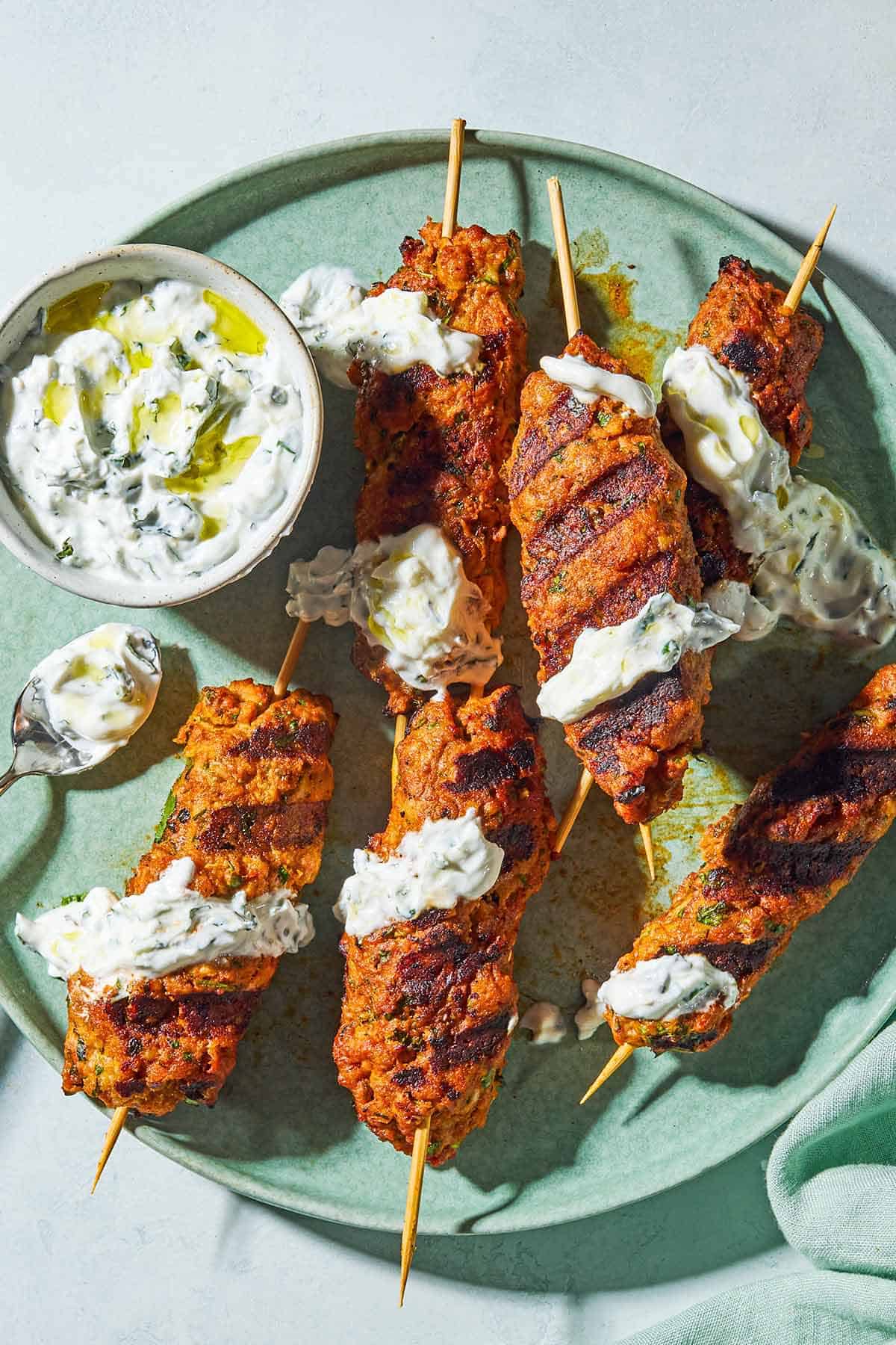 Six grilled chicken kofta kebabs topped with mint yogurt sauce on a plate with the rest of the sauce in a bowl, and spoon.