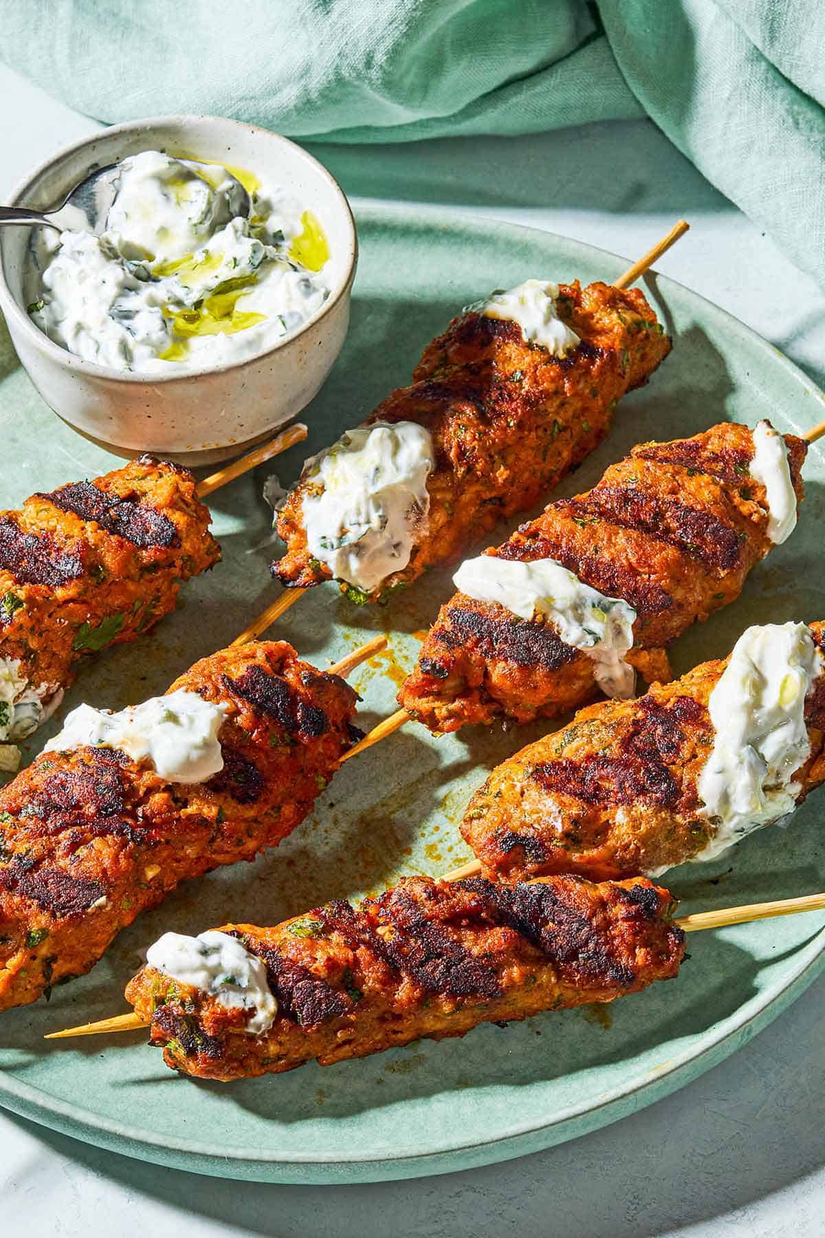 A close up of six grilled chicken kofta kebabs topped with mint yogurt sauce on a plate with the rest of the sauce in a bowl with a spoon.