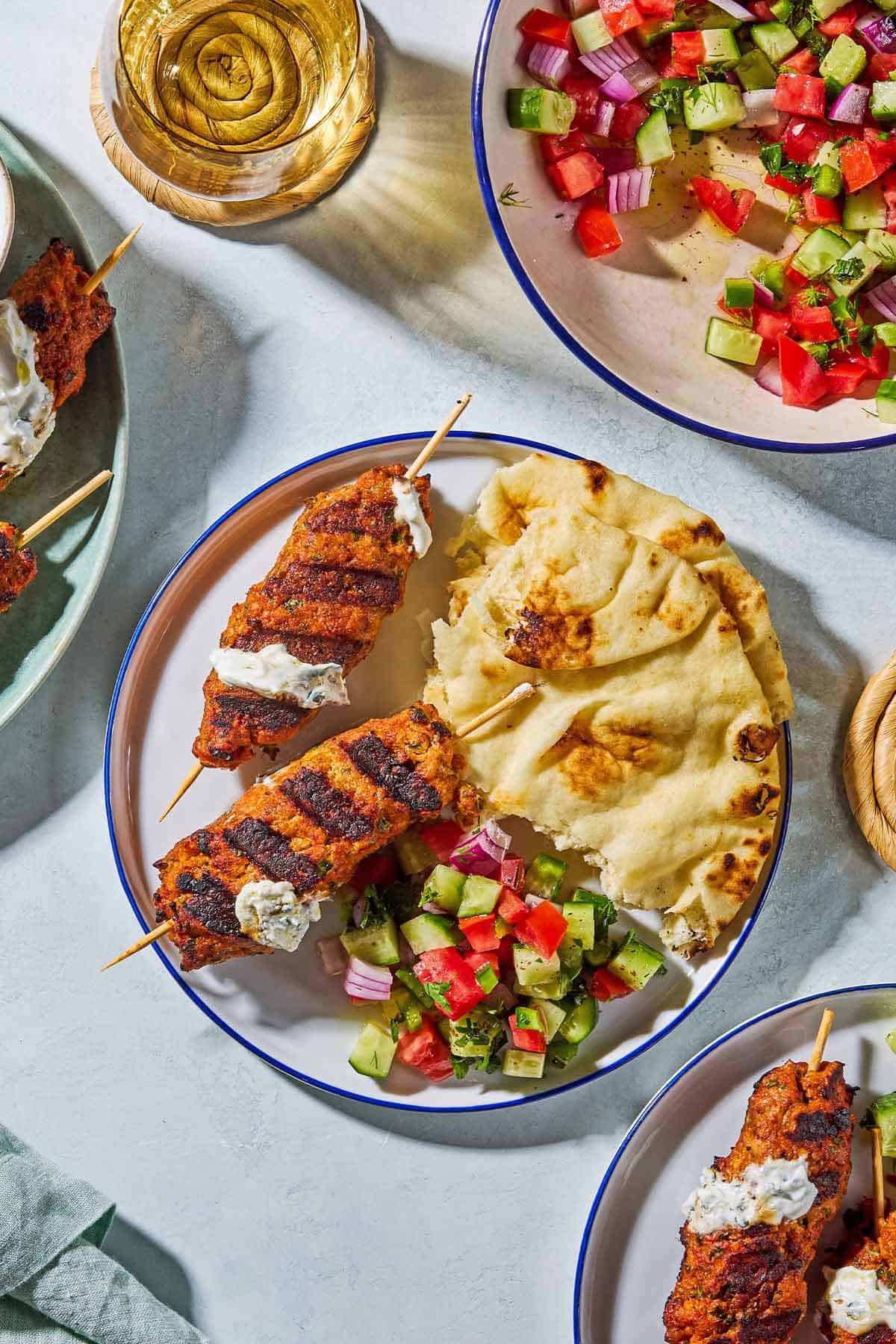 An overhead photo of 2 grilled chicken kofta kebabs topped with mint yogurt sauce on a plate with pieces of pita and a tomato, cucumber salad. Next to this is a bowl of the salad, a glass of water, and 2 more plates with more of the kebabs.